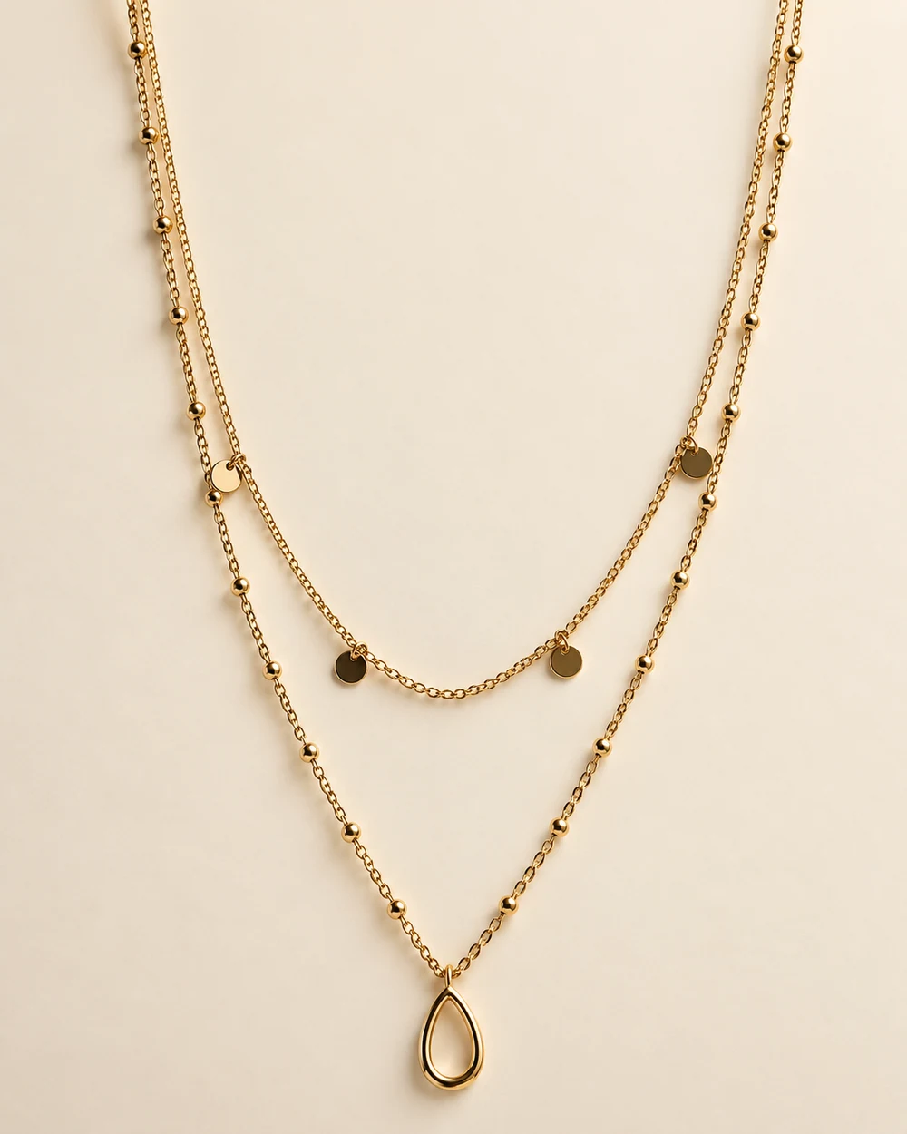 Antoine — Collier à deux niveaux: chaîne fine et petits disques suspendus sur le rang court, chaîne ponctuée de billes et pendentif goutte évidée sur le rang long.
